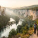 Panorama naturel de l’Ardèche avec une rivière sinueuse, des falaises calcaires, une forêt méditerranéenne et deux randonneurs sur un sentier au lever du soleil.