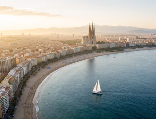 Vue aérienne réaliste de Barcelone au lever du soleil, avec une plage urbaine, un voilier sur la Méditerranée et la silhouette de la Sagrada Família au loin.