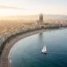 Vue aérienne réaliste de Barcelone au lever du soleil, avec une plage urbaine, un voilier sur la Méditerranée et la silhouette de la Sagrada Família au loin.